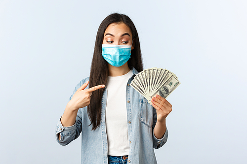 Social distancing lifestyle, covid-19 pandemic business and employement concept. Excited pleased asian girl in medical mask pointing finger at cash, found big money.