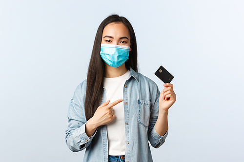 Social distancing lifestyle, covid-19 pandemic and contactless shopping concept. Cheerful cute asian female in medical mask pointing at credit card, advice pay online, order from home.