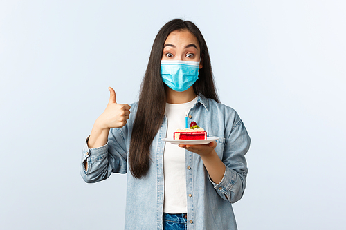 Social distancing lifestyle, covid-19 pandemic, celebrating holidays during coronavirus concept. Happy smiling asian birthday girl in medical mask, show bday cake and thumb-up pleased.