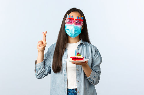 Social distancing lifestyle, covid-19 pandemic, celebrating holidays concept. Hopeful cute asian b-day girl in party glasses and medical mask, holding birthday cake and cross fingers make wish.