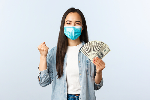 Social distancing lifestyle, covid-19 pandemic business and employement concept. Rejoicing happy asian woman got salary, wear medical mask, fist pump and showing money, triumphing at payday.