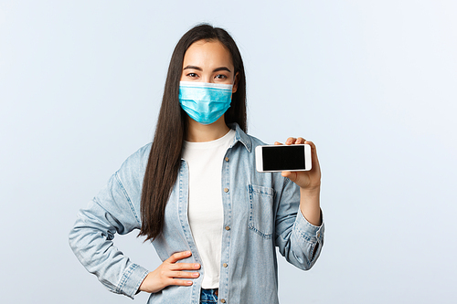 Social distancing lifestyle, covid-19 pandemic and technology concept. Cheerful smiling asian woman in medical mask, showing smartphone display horizontally, promote app.