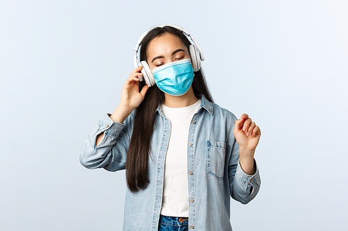 Social distancing lifestyle, covid-19 pandemic and self-isolation leisure concept. Carefree dreamy asian girl in medical mask enjoying listening music in wireless headphones, close eyes relaxed.