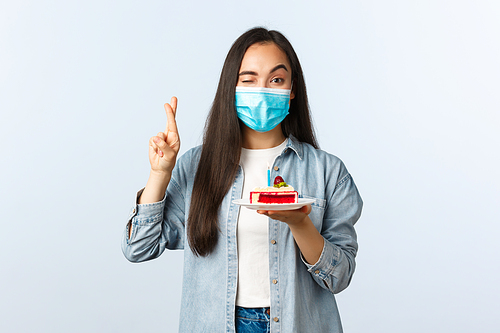 Social distancing lifestyle, covid-19 pandemic, celebrating holidays during coronavirus concept. Hopeful cute asian girl in medical mask, holding birthday cake, cross fingers as making wish.