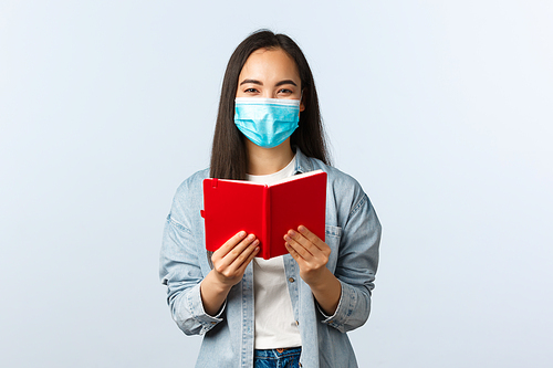 Covid-19 pandemic, education during coronavirus, back to school concept. Cheerful friendly smiling asian woman in medical mask prepare for college exam, studying notes in notebook.