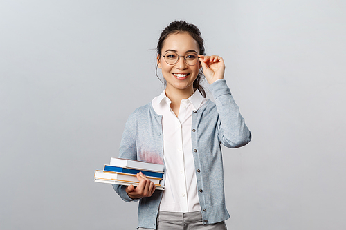 Education, teachers, university and schools concept. Young female student, asian girl studying for exams, fixing glasses on eyes and smiling, carry books to library, stand grey background.