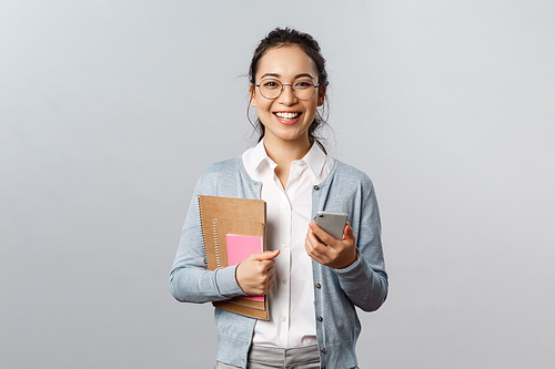 Education, teachers, university and schools concept. Cheerful smart young female tutor, holding smartphone and notebooks with books, prepare lessons, study material, grey background.