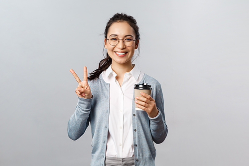 Office lifestyle, business and people concept. Optimistic, friendly-looking asian girl show peace sign, smiling kawaii, enjoying morning fresh cup coffee from local cafe, stand grey background.