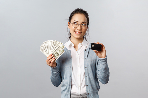 Business, finance and investment concept. Thoughtful smiling young asian rich woman in glasses, bite lip curious, look up while thinking what buy on her prize, hold money dollars and credit card.