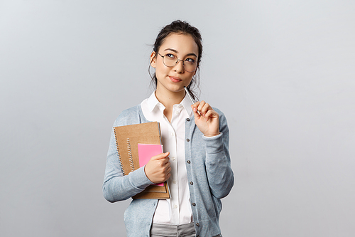 Education, teachers, university and schools concept. Thoughtful asian female student, solving puzzle, look up with intrigued, thinking face, bite pencil while pondering, hold notebooks.