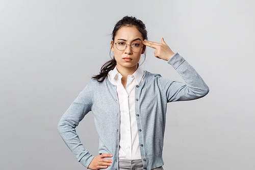 Portrait of sick and tired asian woman hear something stupid, shoot herself with finger gun as a reaction to annoying bothering conversation, staring skeptical camera, standing grey background.