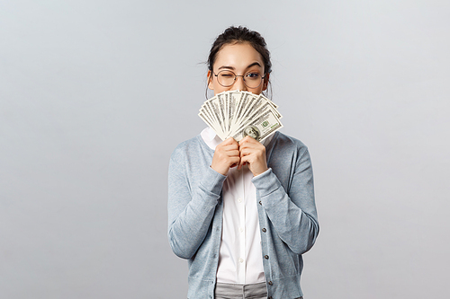 Business, finance and investment concept. Happy, pleased pretty asian woman, rich girl playfully raise eyebrow hiding satisfied smile behind money dollars, win cash prize, grey background.