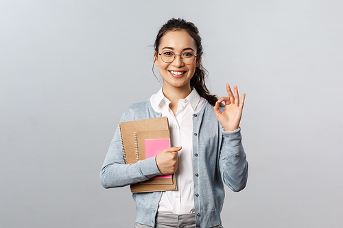 Education, teachers, university and schools concept. Excellent school, join our online lesson. Portrait of satisfied young female student in glasses, show okay sign and hold notebooks with homework.
