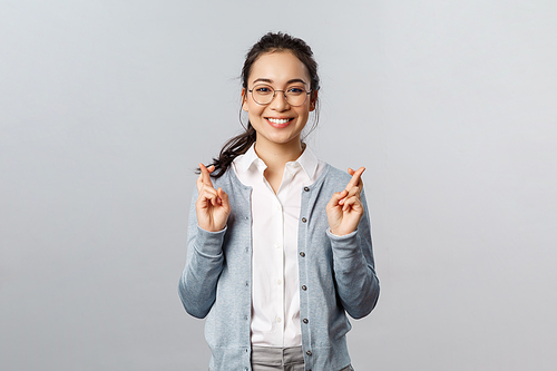 Emotions, people and lifestyle concept. Optimistic happy, smiling asian girl having faith all be alright, cross fingers good luck, hopeful anticipating positive results, pleading over grey background.