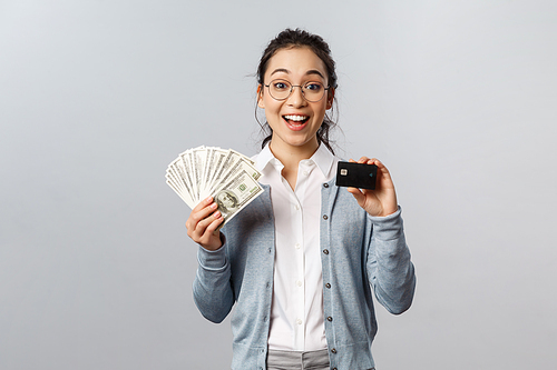 Business, finance and investment concept. Amused happy asian girl decides save for her vacation, place money on deposit, showing cash dollars and credit card, smiling upbeat.