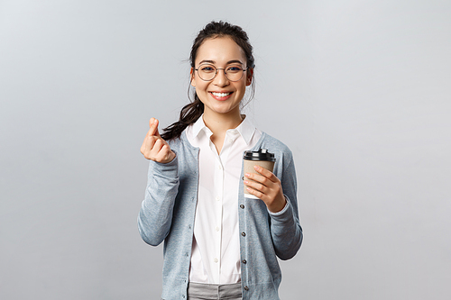 Office lifestyle, business and people concept. Friendly attractive asian female employee, girl showing korean heart with fingers and smiling, drinking take-away coffee, recommend visit cafe.