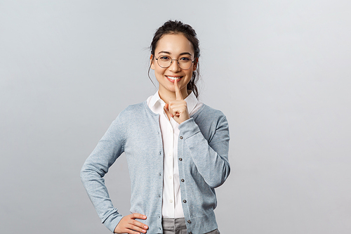 Education, studying and online classes concept. Charming young asian girl in glasses asking polite to keep quiet, shush with smile, hold finger over mouth, be silent please, hold it a secret.
