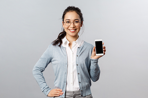 Technology, online and mobile lifestyle concept. Cheerful asian woman in glasses introduce new application, internet store link, showing smartphone display with pleased smile.