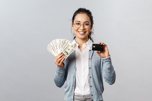 Business, finance and investment concept. Young friendly-looking female employee in glasses, asian woman holding cash and credit card, show different ways of paying online or offline purchase.