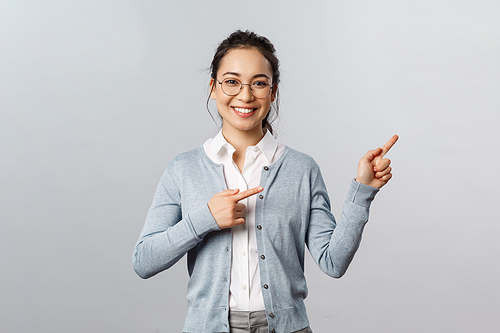 Education, studying and online classes concept. Cheerful asian woman, teacher pointing fingers right at presentation or chart, smiling friendly, helping find way, showing promo, grey background.