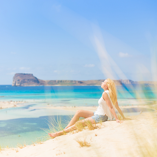 Relaxed woman enjoying sun, freedom and life an a beautiful sandy beach of Balos in Greece. Young lady feeling free, relaxed and happy. Vacations, freedom, happiness, enjoyment and well being.