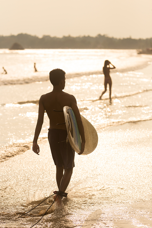 Silhouette of unrecognizable male surfer walking at tropical beach of Midigama, Sri Lanka at sunsen with surf board in his hands.