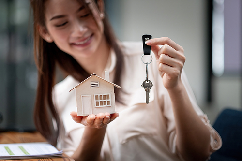 mortgage, real estate and property concept - close up of hands holding house model and home keys at modern office.