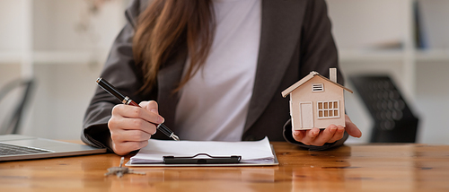 Close up businesswoman realtor taking notes and holding house model, sitting at desk with paper house model and keys, female real estate agent manager or broker making deal, preparing documents, mortgage and property