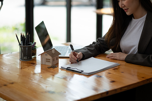 Crop close up house model miniature on realtor table, businesswoman real estate agent broker writing with documents, taking notes, preparing contract for deal with client, mortgage or rent concept.