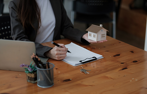 Crop close up house model miniature on realtor table, businesswoman real estate agent broker writing with documents, taking notes, preparing contract for deal with client, mortgage or rent concept.