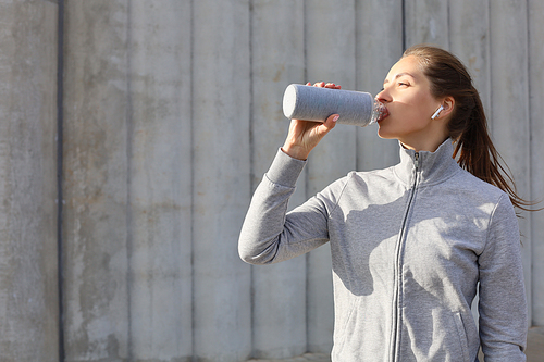 Beautiful female runner standing outdoors, drinking water from bottle. Fitness woman takes a break after running workout.