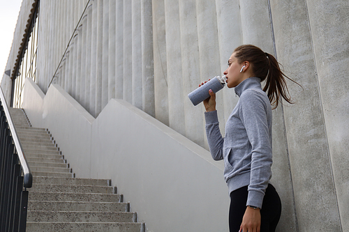 Beautiful female runner standing outdoors, drinking water from bottle. Fitness woman takes a break after running workout.