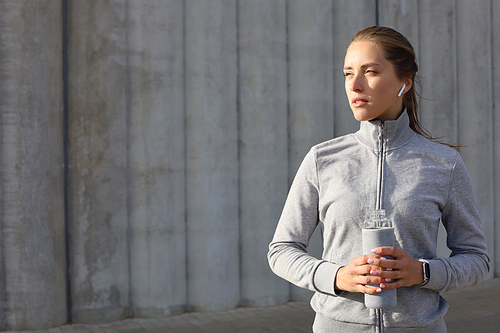 Beautiful female runner is standing outdoors holding water bottle. Fitness woman takes a break after running workout.