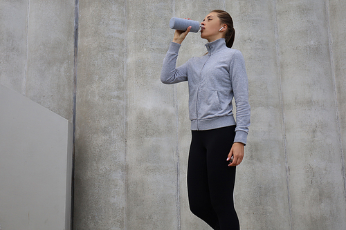 Beautiful female runner standing outdoors, drinking water from bottle. Fitness woman takes a break after running workout.