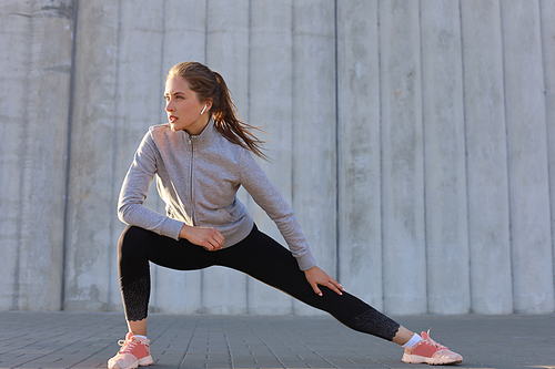 Young attractive sporty girl warming up outdoors, doing stretching at sunset or sunrise in city.