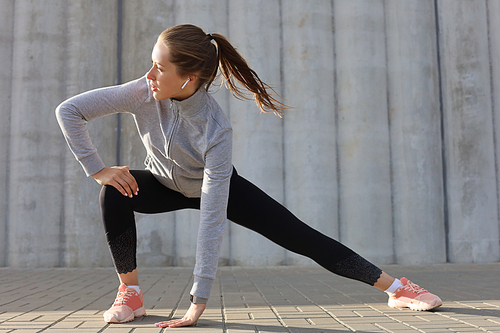 Young attractive sporty girl warming up outdoors, doing stretching at sunset or sunrise in city.