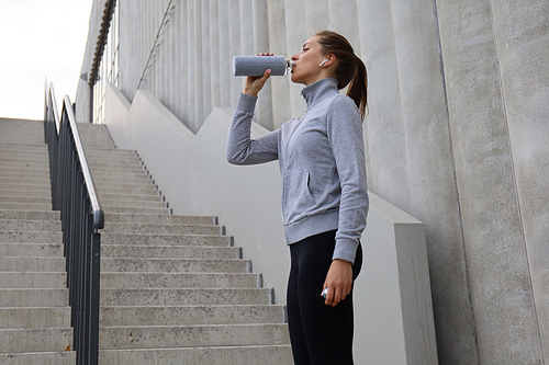 Beautiful female runner standing outdoors, drinking water from bottle. Fitness woman takes a break after running workout.