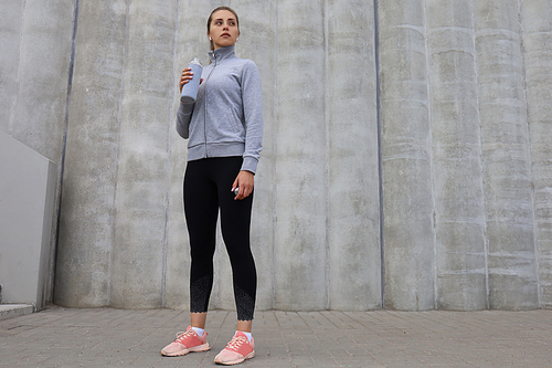 Beautiful female runner is standing outdoors holding water bottle. Fitness woman takes a break after running workout.