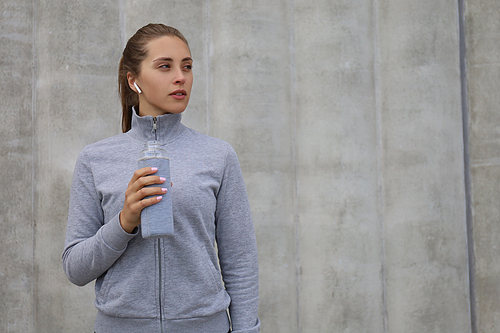 Beautiful female runner is standing outdoors holding water bottle. Fitness woman takes a break after running workout.