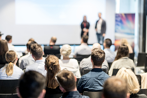 Business and entrepreneurship symposium. Speaker giving a talk at business meeting. Audience in conference hall. Rear view of unrecognized participant in audience.