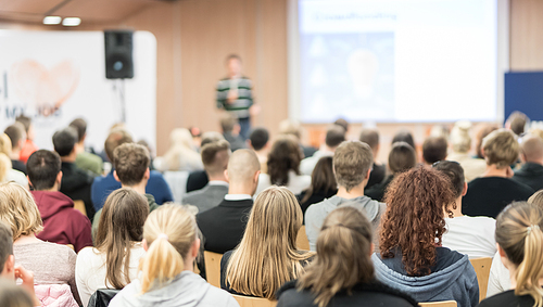 Speaker giving presentation in lecture hall at university. Participants listening to lecture and making notes.