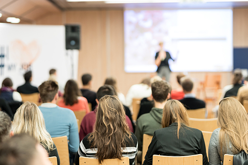 Business and entrepreneurship symposium. Female speaker giving a talk at business meeting. Audience in conference hall. Rear view of unrecognized participant in audience.