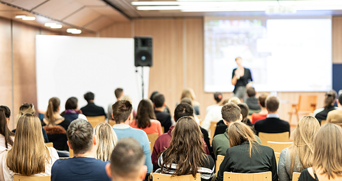 Business and entrepreneurship symposium. Female speaker giving a talk at business meeting. Audience in conference hall. Rear view of unrecognized participant in audience.