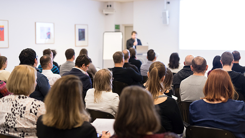 Business and entrepreneurship symposium. Female speaker giving a talk at business meeting. Audience in conference hall. Rear view of unrecognized participant in audience. Copy space on whitescreen.