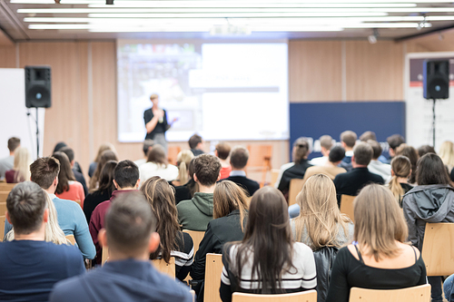 Business and entrepreneurship symposium. Female speaker giving a talk at business meeting. Audience in conference hall. Rear view of unrecognized participant in audience.