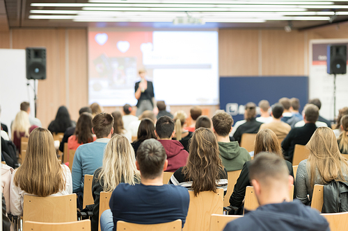 Business and entrepreneurship symposium. Female speaker giving a talk at business meeting. Audience in conference hall. Rear view of unrecognized participant in audience.