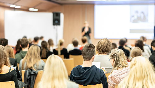 Business and entrepreneurship symposium. Female speaker giving a talk at business meeting. Audience in conference hall. Rear view of unrecognized participant in audience.