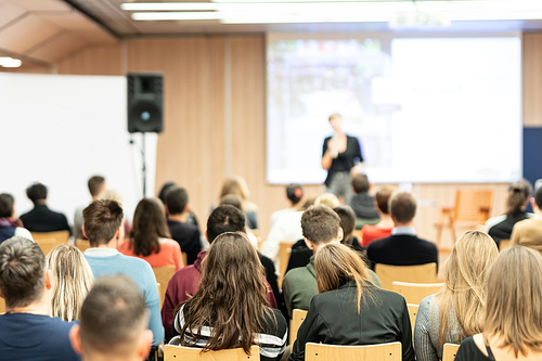 Business and entrepreneurship symposium. Female speaker giving a talk at business meeting. Audience in conference hall. Rear view of unrecognized participant in audience.