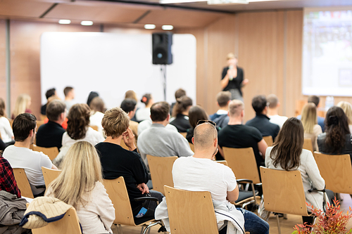 Business and entrepreneurship symposium. Female speaker giving a talk at business meeting. Audience in conference hall. Rear view of unrecognized participant in audience.