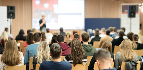 Business and entrepreneurship symposium. Female speaker giving a talk at business meeting. Audience in conference hall. Rear view of unrecognized participant in audience.